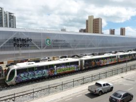 Veículo leve sobre trilhos é visto de lado, parado na estação Papicu, em Fortaleza. O céu, ao fundo, tem muitas nuvens, embora o dia esteja ensolarado. O veículo é branco com alguns grafites ao longo de sua extensão. Mais abaixo na imagem, pode-se ver a rua paralela a estação com veículos circulando.