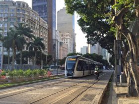 VLT carioca visto no entardecer do Rio de Janeiro, circulando na Cinelândia. Ao fundo, do lado esquerdo na diagonal, se vê os prédios da região. Do lado direito, as árvoes, Ao lado do trem, do lado direito está um motoqueiro circulando com o veículo.