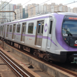 Frente do trem é vista de frente, em movimento circulando na linha lilas. O trem é cinza metálico e lilás. Foto diurna.