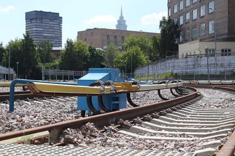 Terceiro trilho (com cobertura amarela), ao lado esquerdo dos trilhos, no sistema ferroviário próximo a estação Sokol, em Moscou, Rússia. Ao fundo, vê-se um céu azul claro e edifícios da cidade.