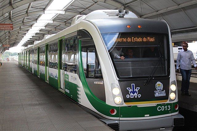 VLT em operação no Metrô de Teresina durante o dia, parado em uma estação com as portas fechadas. Na imagem, o trem é visto na diagonal, de frente. O veículo é branco com detalhes em verde. A estação tem teto em formato circular com uma abertura no alto que permite entrada de luz natural. Há uma pessoa de pé, do lado direito da imagem vestindo camisa clara e calça jeans. No letreiro do trem, pode-se ler "Governo do Estado do Piauí".